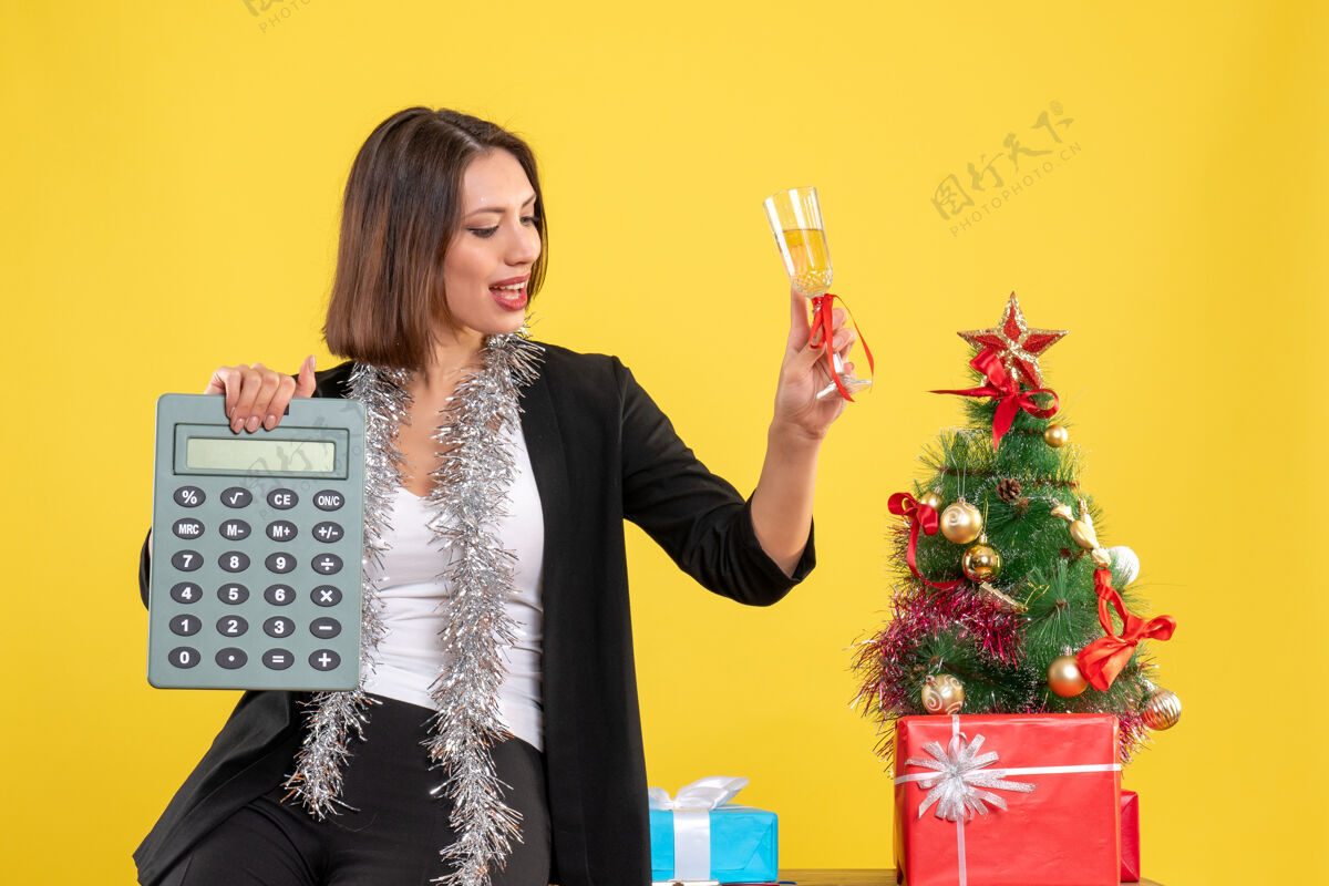 人圣诞气氛积极美丽的女士站在办公室里 拿着计算器在黄色的办公室里举酒微笑酒成人