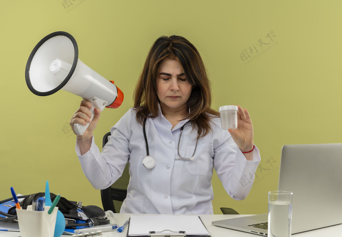 人中年女医生穿着医用长袍和听诊器坐在办公桌旁 手持医疗工具和笔记本电脑 手持扬声器和医用烧杯 闭眼隔离桌子绿色工具