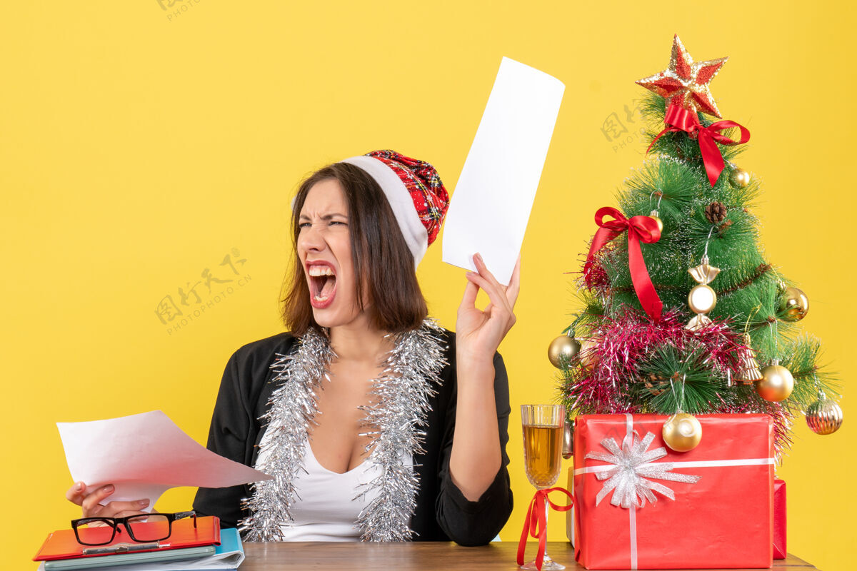 快乐穿着西装 戴着圣诞老人帽和新年装饰品的商务女士感到紧张 坐在办公室的桌子旁 桌上放着圣诞树桌子紧张套装