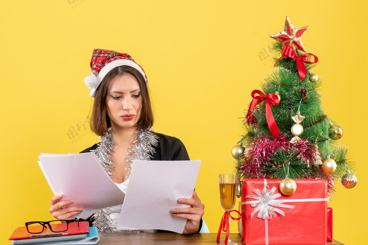 桌子穿着西装 戴着圣诞老人帽和新年装饰品的商务女士正在检查文件 坐在办公室里一张桌子旁 桌子上放着圣诞树坐检查帽子