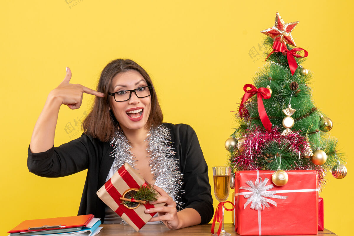 快乐一位穿着西装戴着眼镜的商务女士手里拿着礼物 坐在一张桌子旁 桌子上放着一棵圣诞树人新郎抱着