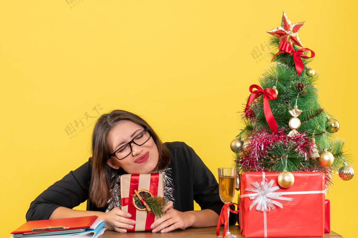 快乐一位穿着西装 戴着眼镜的商务女士手里拿着礼物 梦想着坐在办公室的桌子旁 桌上放着一棵圣诞树西装花束树