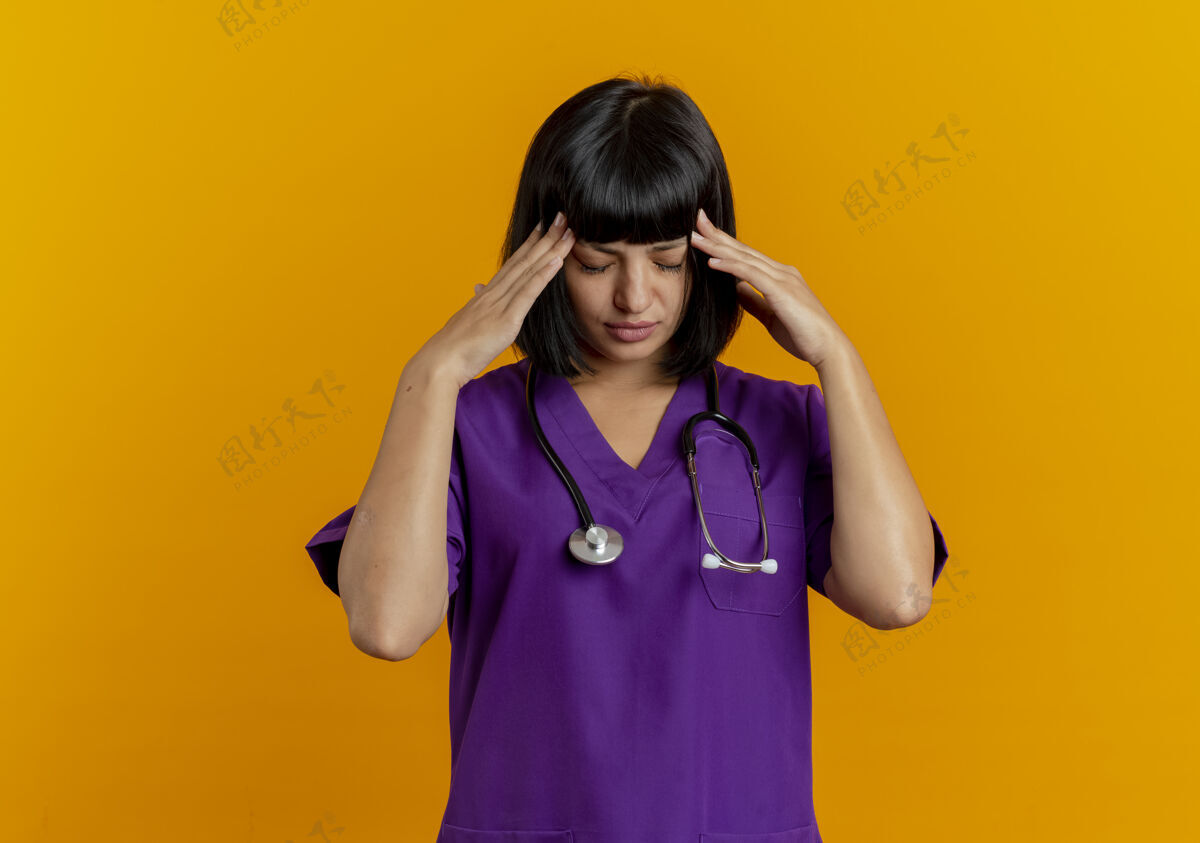 太阳穴恼怒的年轻黑发女医生穿着制服 用听诊器将手放在隔离在橙色背景上的太阳穴上 并留有复印空间副本年轻太空