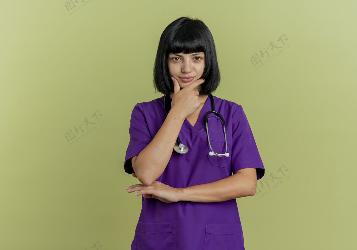 女性自信的年轻黑发女医生穿着制服 用听诊器将手放在下巴上 隔离在橄榄绿背景上 留有复印空间年轻复制自信