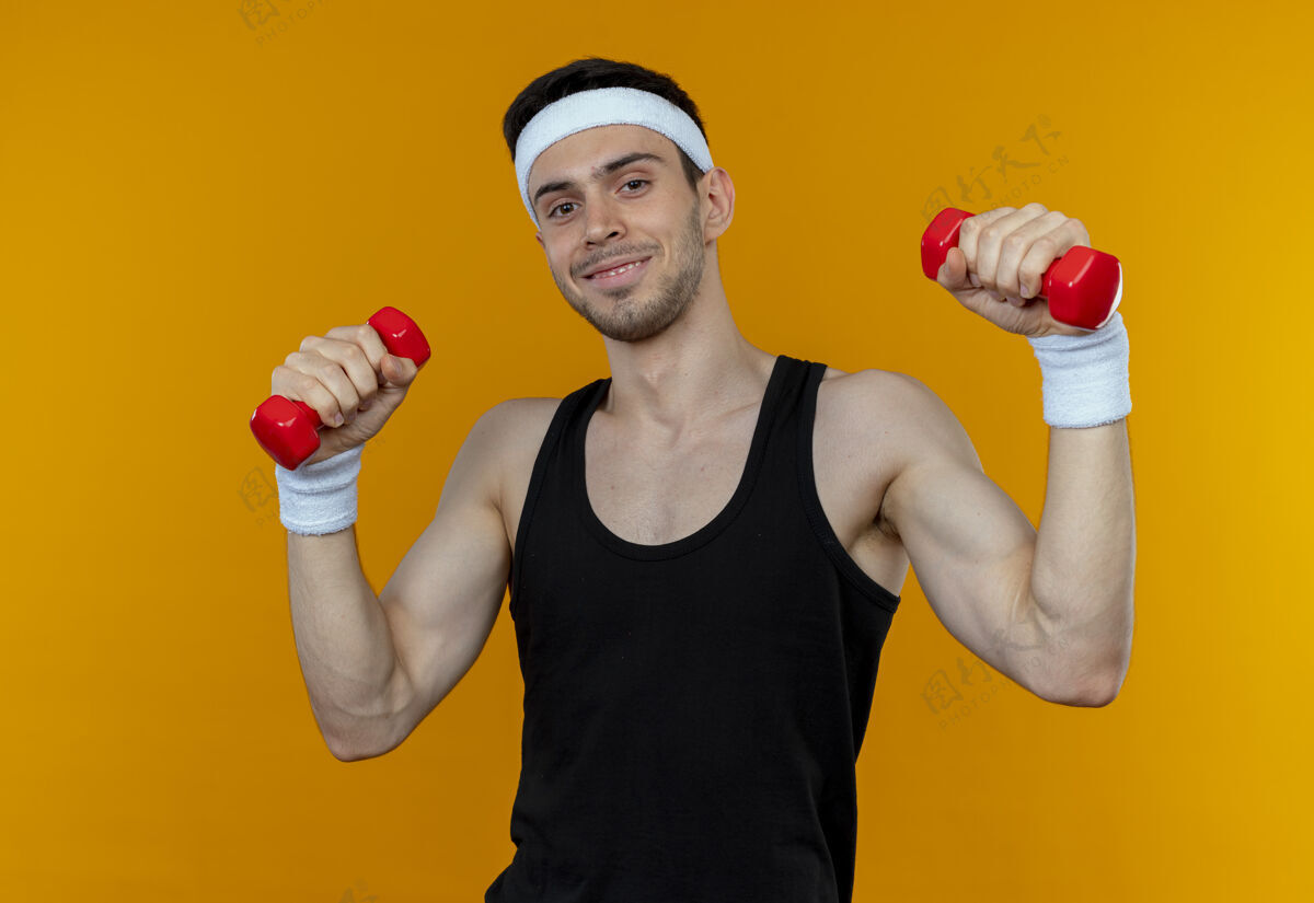 运动戴着头巾的年轻运动型男子举着哑铃 站在橙色背景下微笑着看着摄像机头带微笑相机