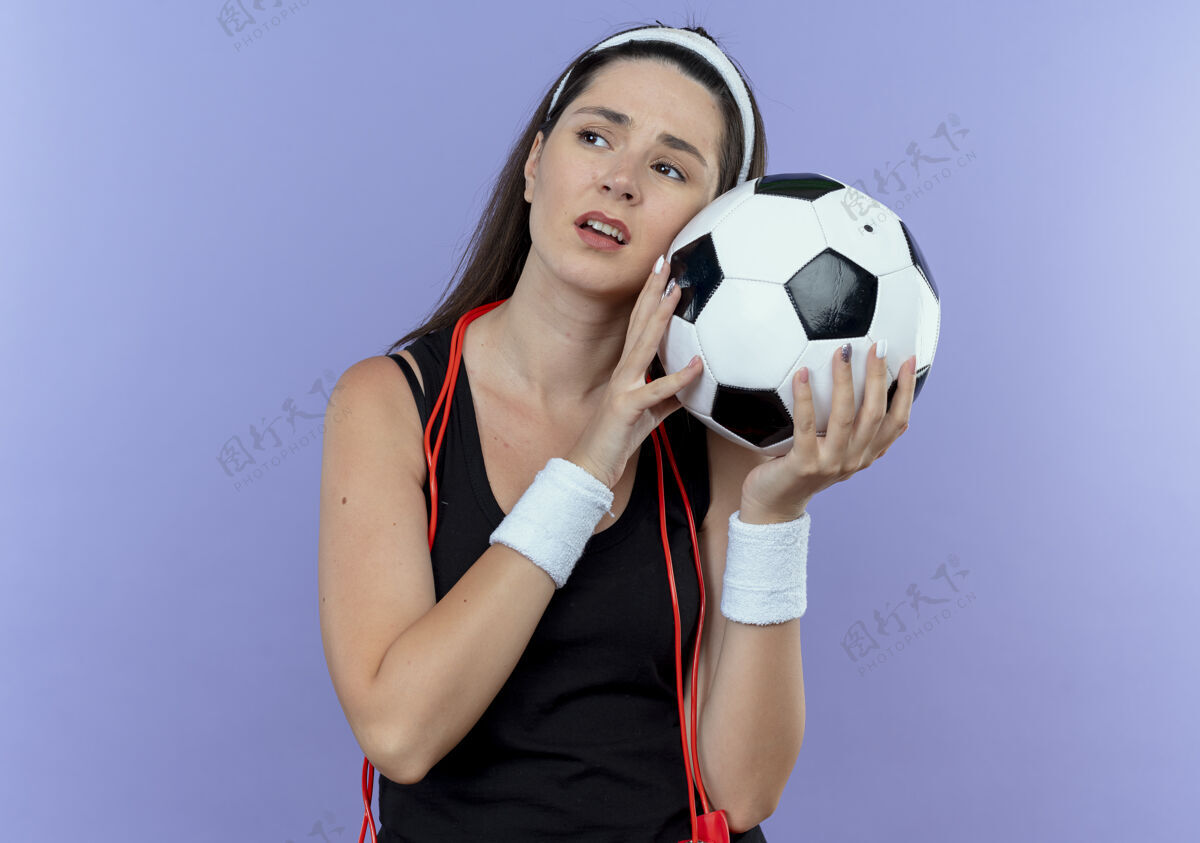 抱着戴着头巾 脖子上系着跳绳 手里拿着足球 看着轮胎 无聊地站在蓝色背景上的年轻健身女士脖子绳子周围