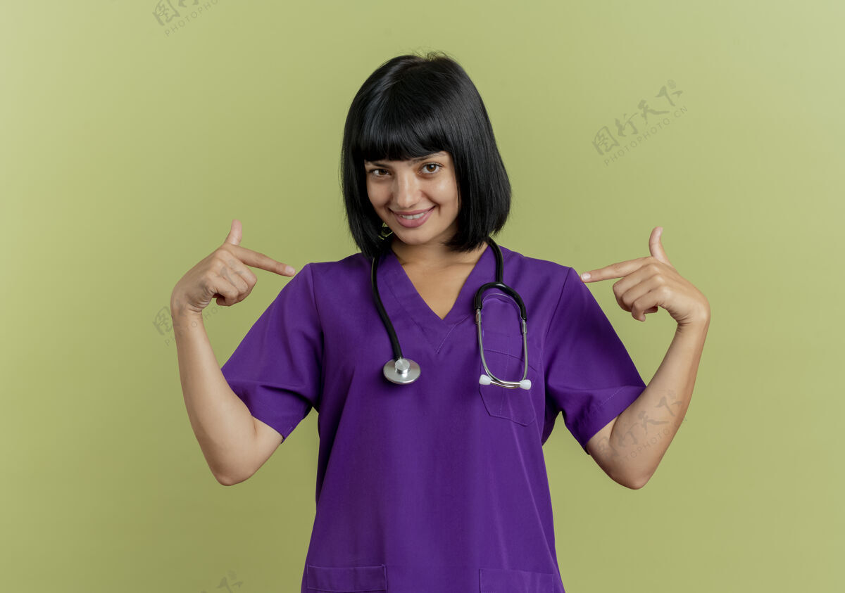 医生微笑着的年轻黑发女医生穿着制服 用听诊器指着自己 两只手孤立地放在橄榄绿的背景上 留有复印空间黑发她自己空间