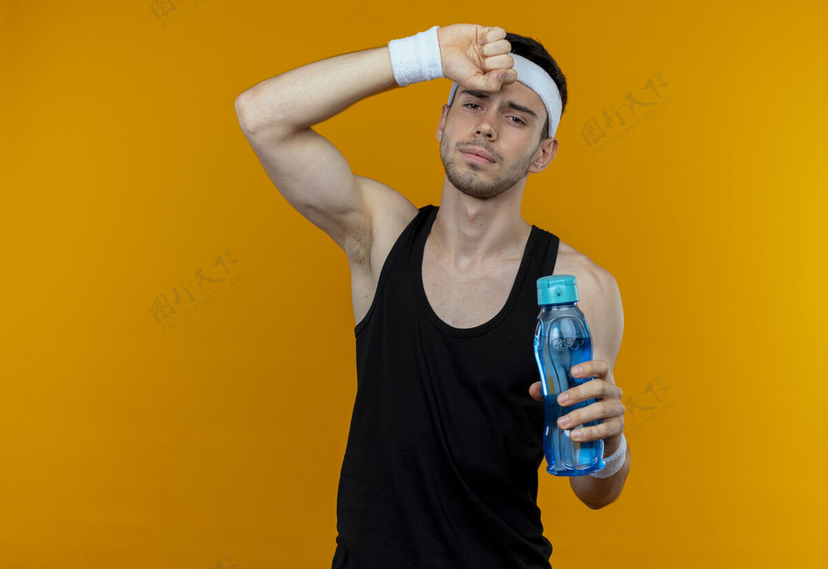 头带站在橙色的背景下 戴着头巾 拿着一瓶水 看上去筋疲力尽的年轻运动型男子筋疲力尽后站着