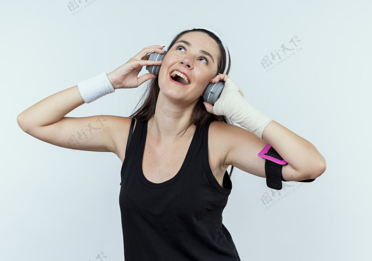 健身戴着头戴式耳机和智能手机臂章的年轻健身女士站在白色背景下欣赏她最喜爱的音乐年轻臂带头带