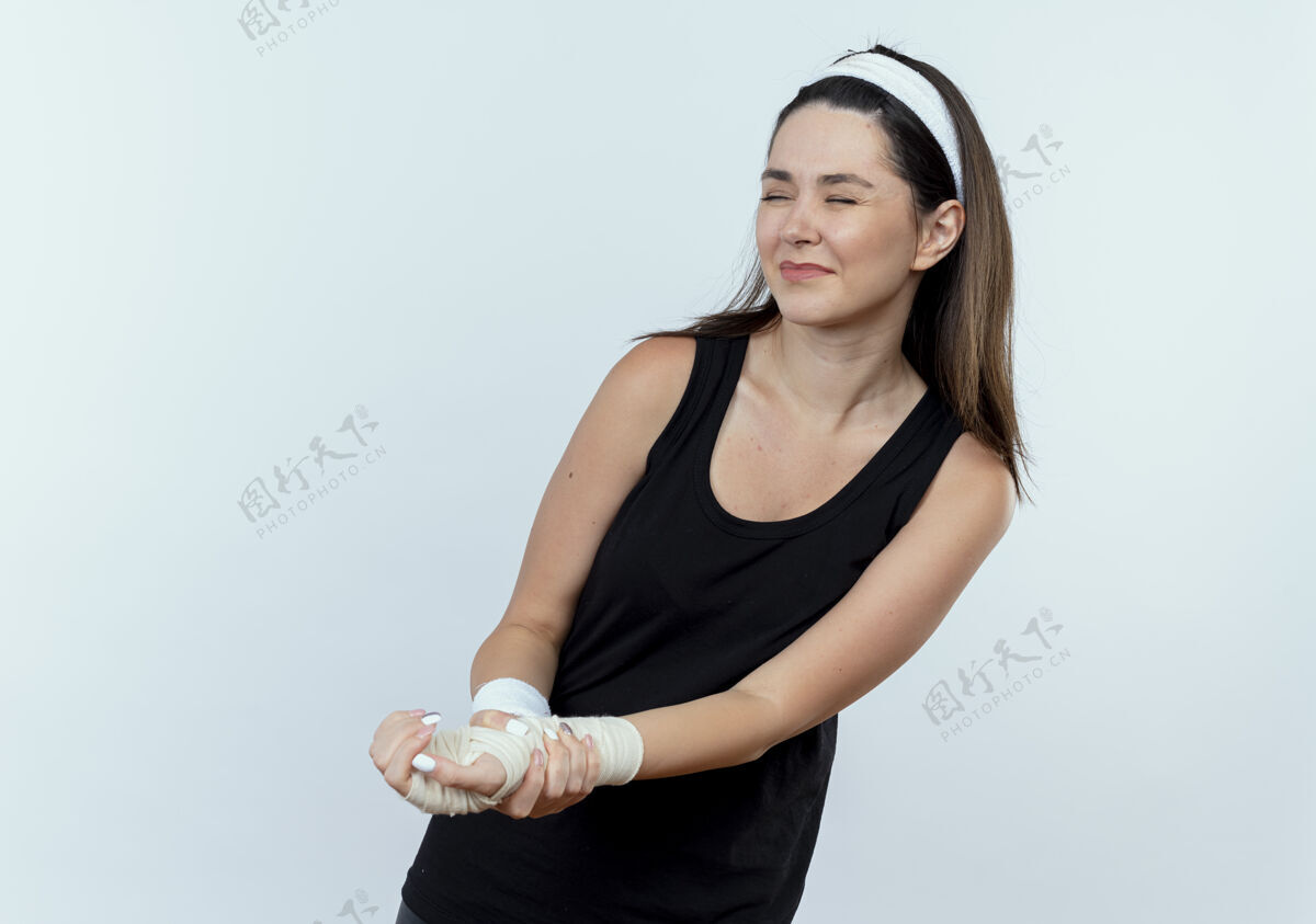 年轻戴着头巾的年轻健身女士触摸着缠着绷带的手腕 站在白色背景上感到疼痛疼痛绷带手腕