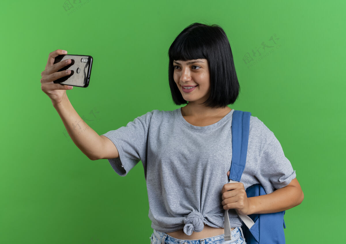 绿色带着微笑的年轻黑发白人女孩背着背包看着手机 在绿色背景下单独拍照 还有复印空间微笑复制空间