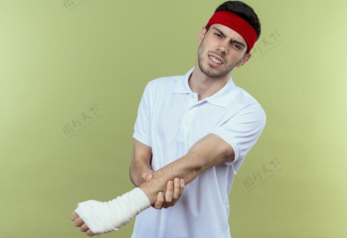 运动戴着头巾的年轻运动型男子站在绿色背景上 摸着缠着绷带的手感到疼痛绷带头带触摸