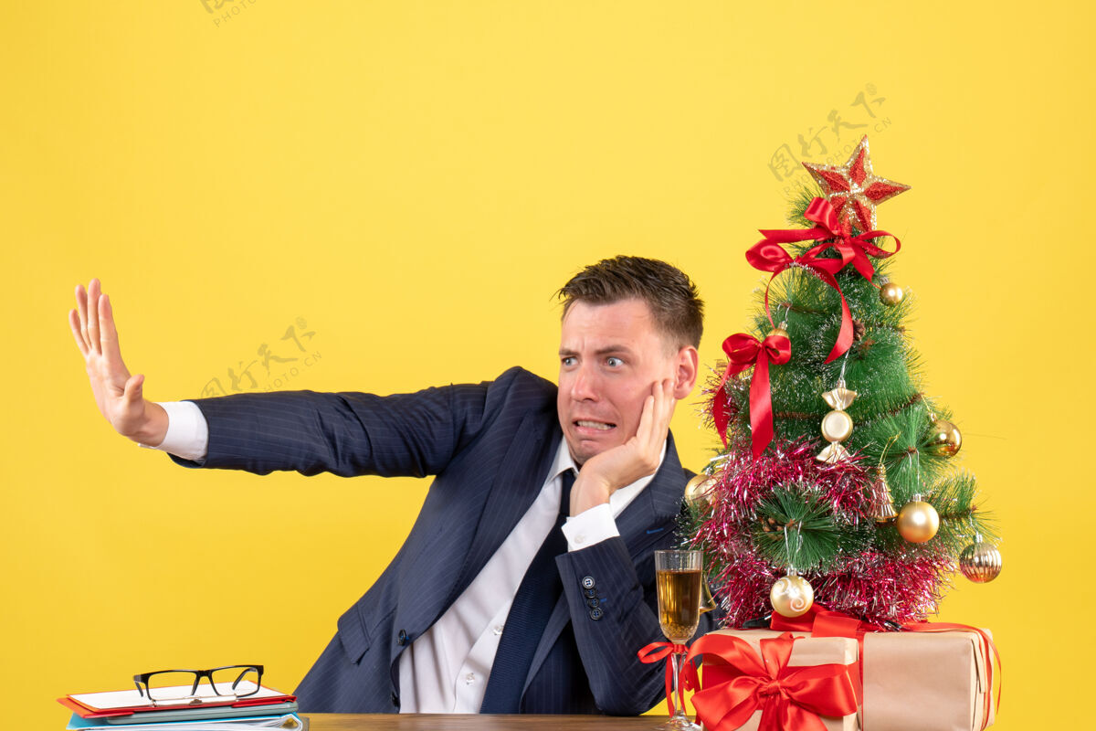 礼物前视图激动的男子试图阻止坐在圣诞树和黄色背景上的礼物附近的桌子上的东西新郎办公室背景
