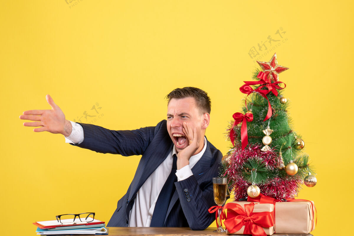 生意人前视图愤怒的男子一边大喊大叫 一边坐在圣诞树和黄色背景上的礼物旁边的桌子上圣诞节坐着办公室