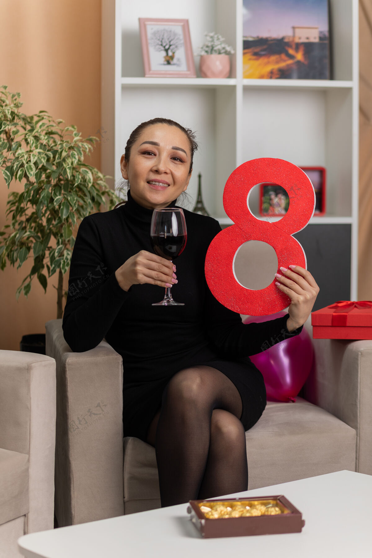 年轻快乐的亚洲女人穿着黑色连衣裙坐在椅子上 手里拿着一杯葡萄酒 手里拿着8号 在灯光明亮的客厅里欢快地笑着 庆祝国际妇女节三月生活椅子微笑