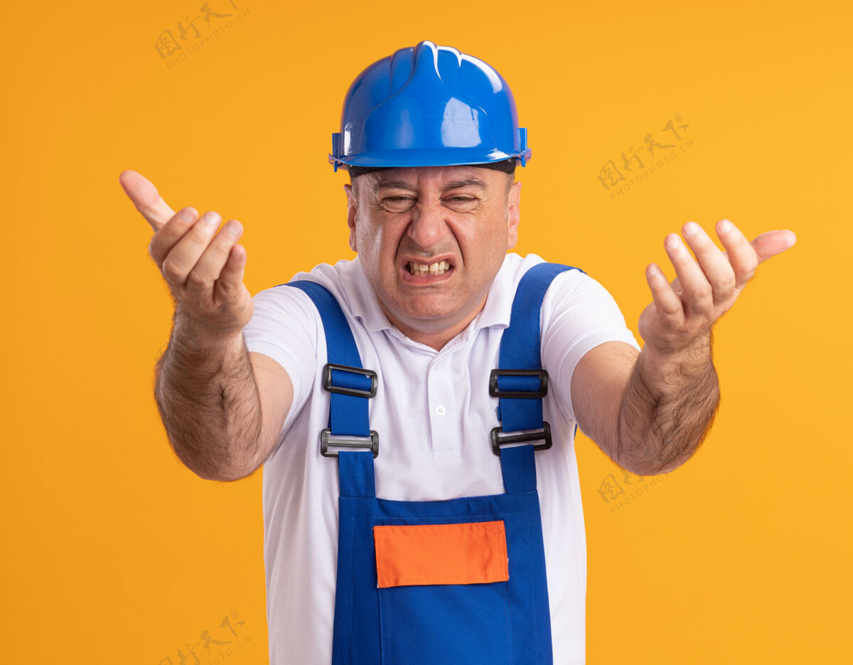 立场恼怒的穿制服的成年建筑工人站在前面 两只手被隔离在橙色的墙上公民姿势衣服
