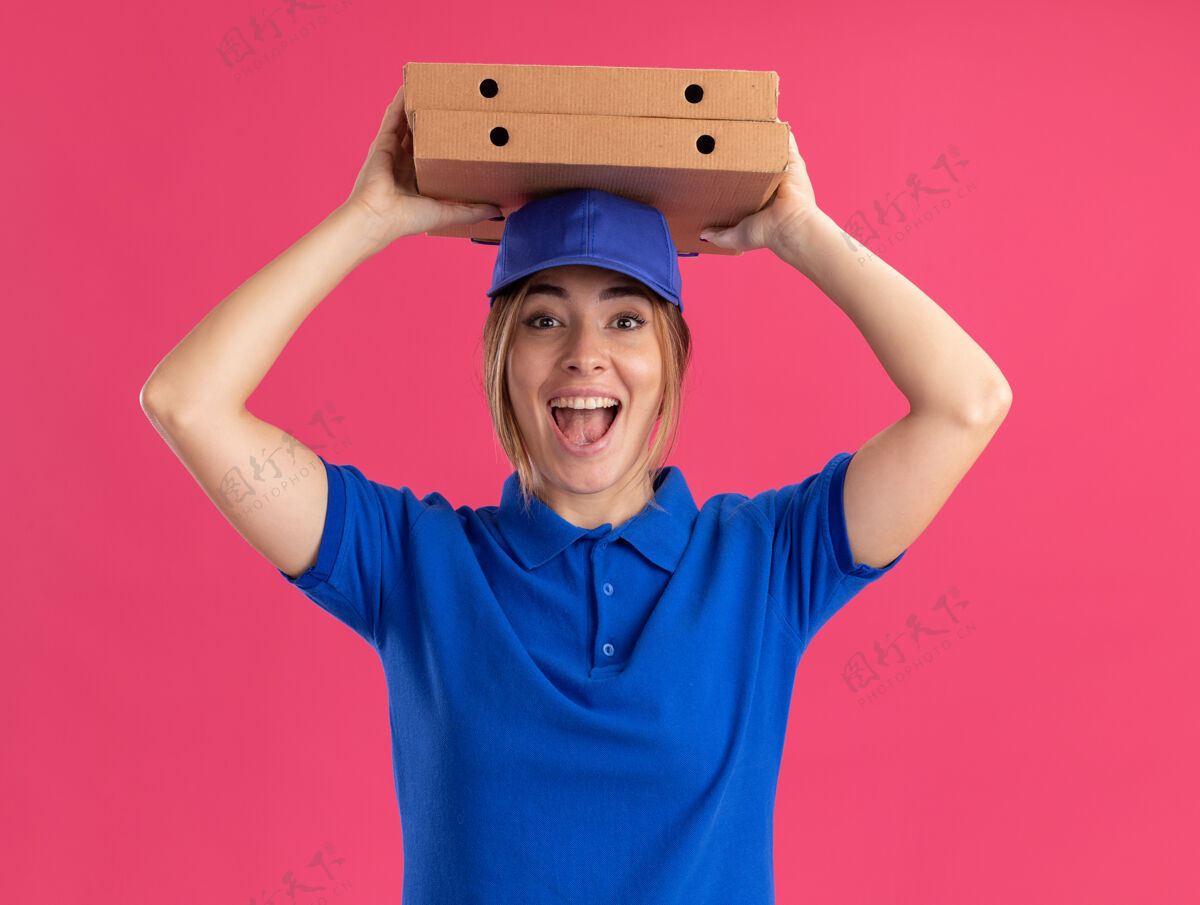 市民穿着制服的年轻漂亮的送货员兴奋地把披萨盒举过头顶 隔离在粉红色的墙上盒子感觉衣服