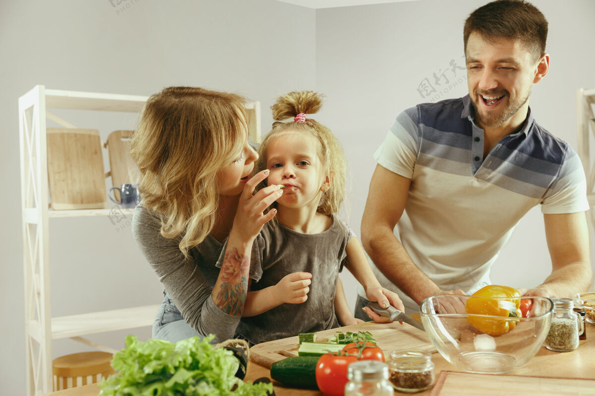 烹饪可爱的小女孩和她美丽的父母正在家里的厨房里切菜 一边微笑着做沙拉家庭生活理念乐趣情绪女性