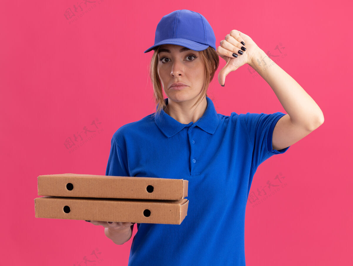 人失望的年轻漂亮的女送货员穿着制服 大拇指朝下 拿着比萨饼盒隔离在粉红色的墙上感觉人年轻人
