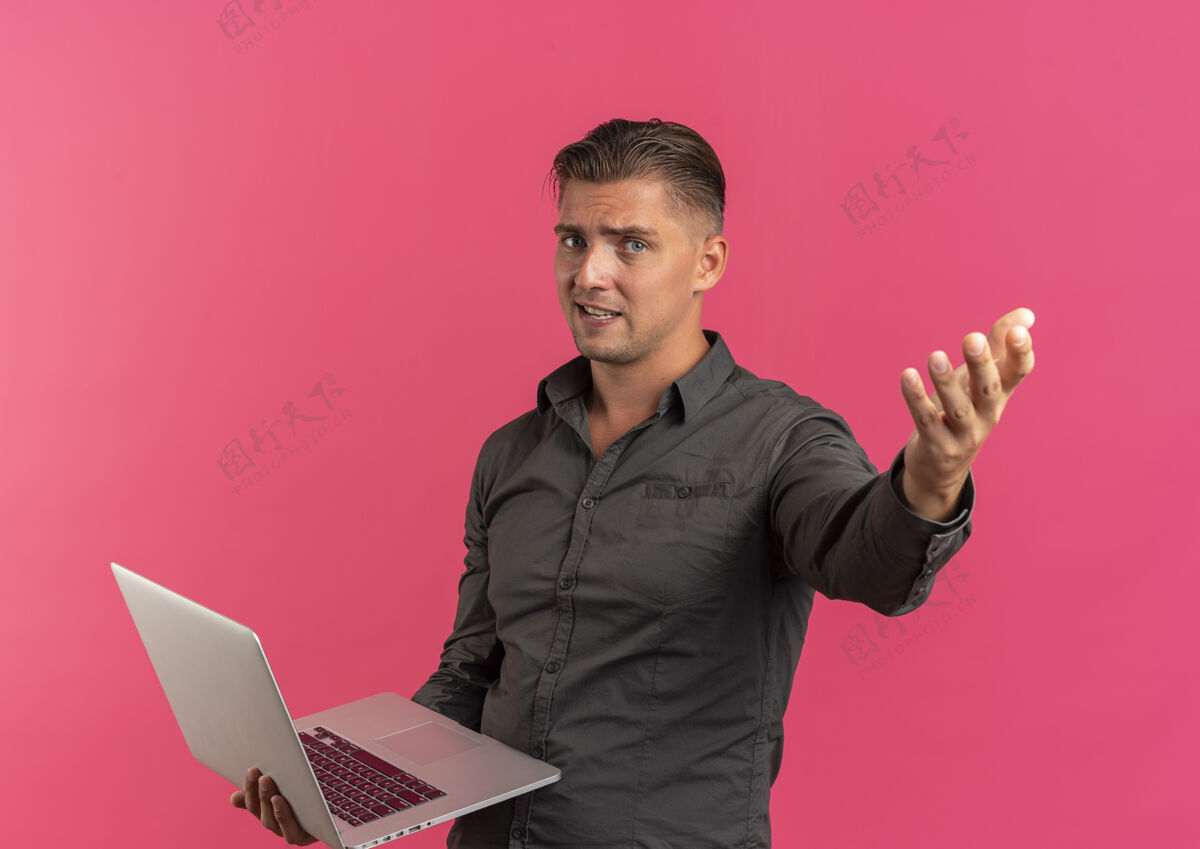 恼怒年轻恼怒的金发帅哥拿着笔记本电脑 在粉色背景上孤立地伸出手来 还有复印空间笔记本电脑英俊拿着