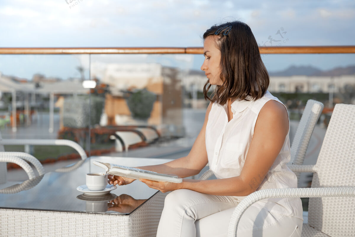 酒店年轻的黑发女人手里拿着一杯热饮和一本书享受着早晨的时光阅读咖啡女士