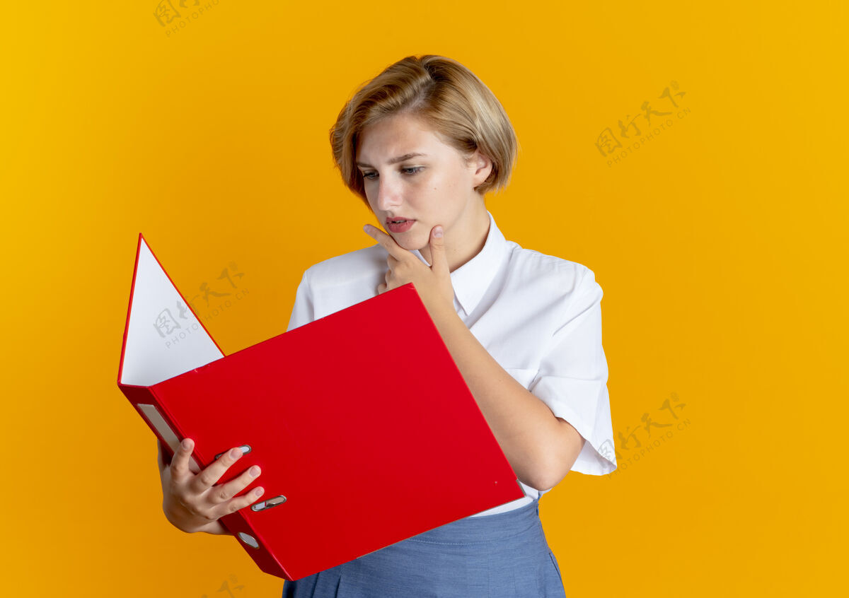 女孩年轻严肃的金发俄罗斯女孩把手放在下巴上看文件夹孤立在橙色背景与复制空间手下巴认真