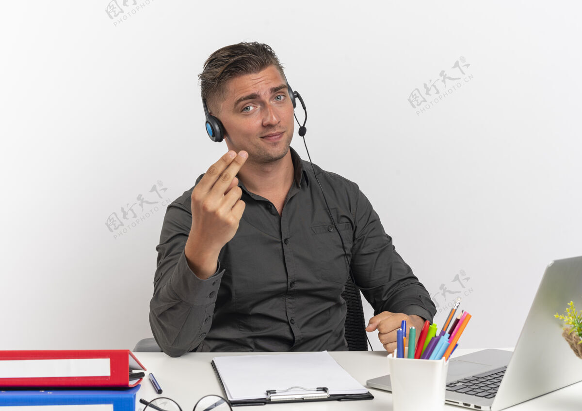 办公室年轻自信的金发上班族戴着耳机坐在办公桌旁 拿着办公工具 用笔记本电脑和手势来到这里 在白色背景下 有复印空间办公桌用工具