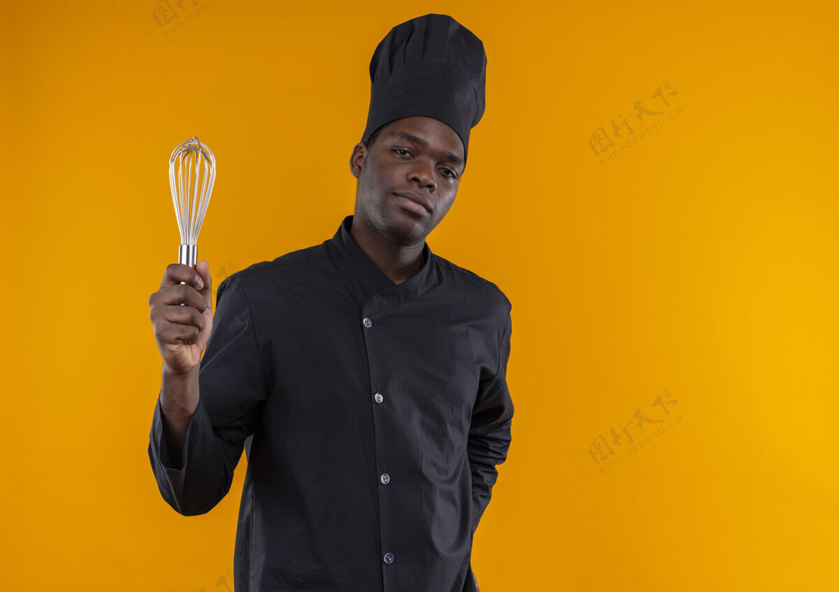 持有年轻自信的美国黑人厨师身着厨师制服 手持惠斯克 看着橙色背景上的相机 并留有复印空间威士忌相机烹饪