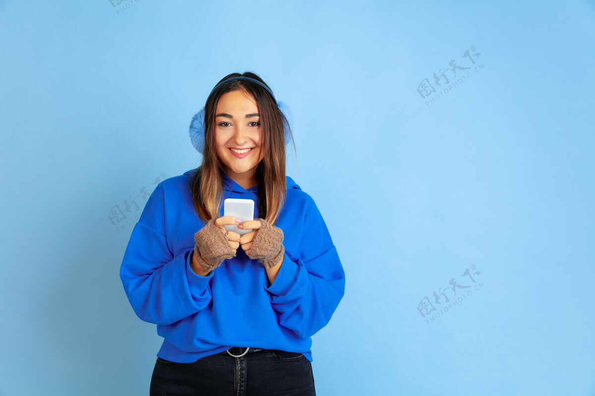 手势使用电话 快乐蓝色工作室背景上的白种女人肖像穿着暖和衣服的漂亮女模特人类情感的概念 面部表情 销售 广告冬天的心情 假期员工执行人
