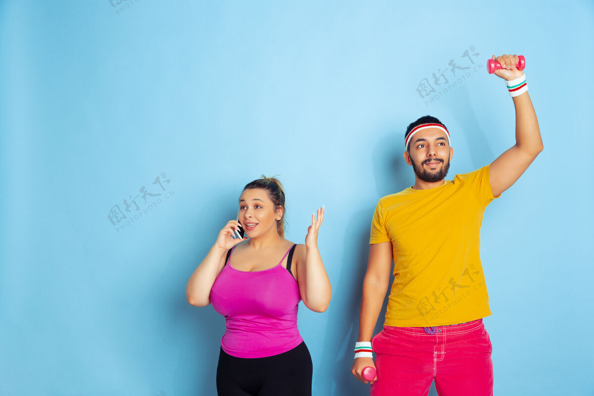交叉年轻漂亮的白人夫妇穿着鲜艳的衣服 在蓝色背景下训练运动概念 人类情感 表达 健康的生活方式 关系 家庭他在训练 她在打电话情侣积极的运动鞋
