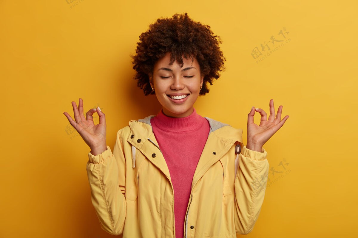 姿势专注开朗的卷发女人双手保持良好的姿势 在室内冥想 闭上眼睛 穿着黄色的休闲服夹克牙关手