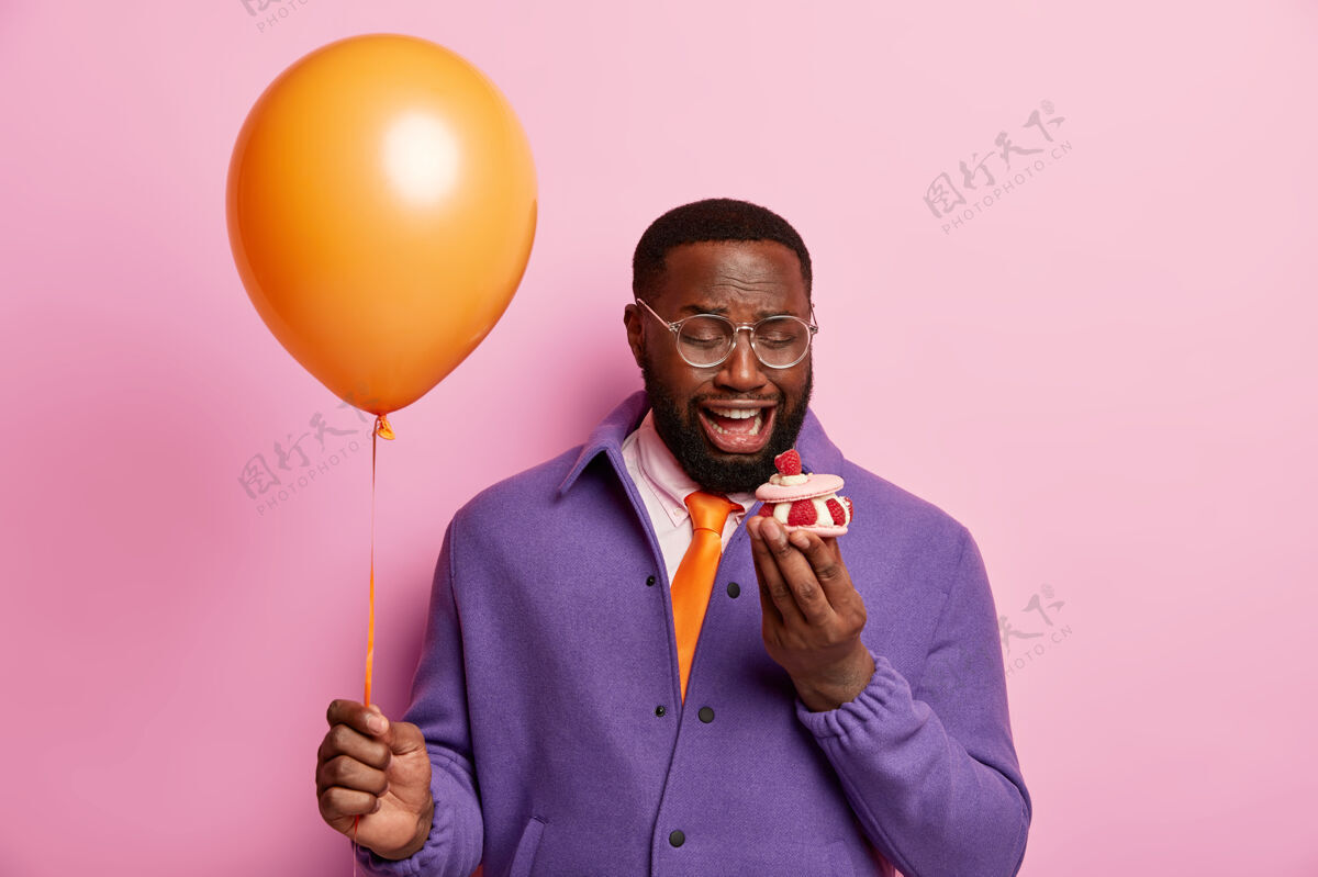 时尚孤独的非洲男人不满地看着小甜纸杯蛋糕 独自庆祝节日 手持气球不快乐墙壁眼镜