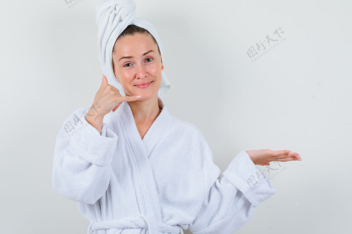 亚洲人身着白色浴衣 毛巾的年轻女士展示着电话手势 看上去很高兴 正前方的景色人女士自然