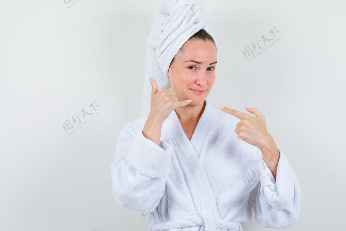新鲜年轻女士指着电话牌 穿着白色浴衣 拿着毛巾 看上去很高兴正视图电话女性浴袍