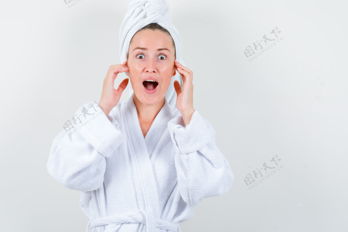 女士年轻的女士手拉着耳朵 穿着白色浴衣 脸上挂着毛巾 脸上露出惊讶的表情手头发模特