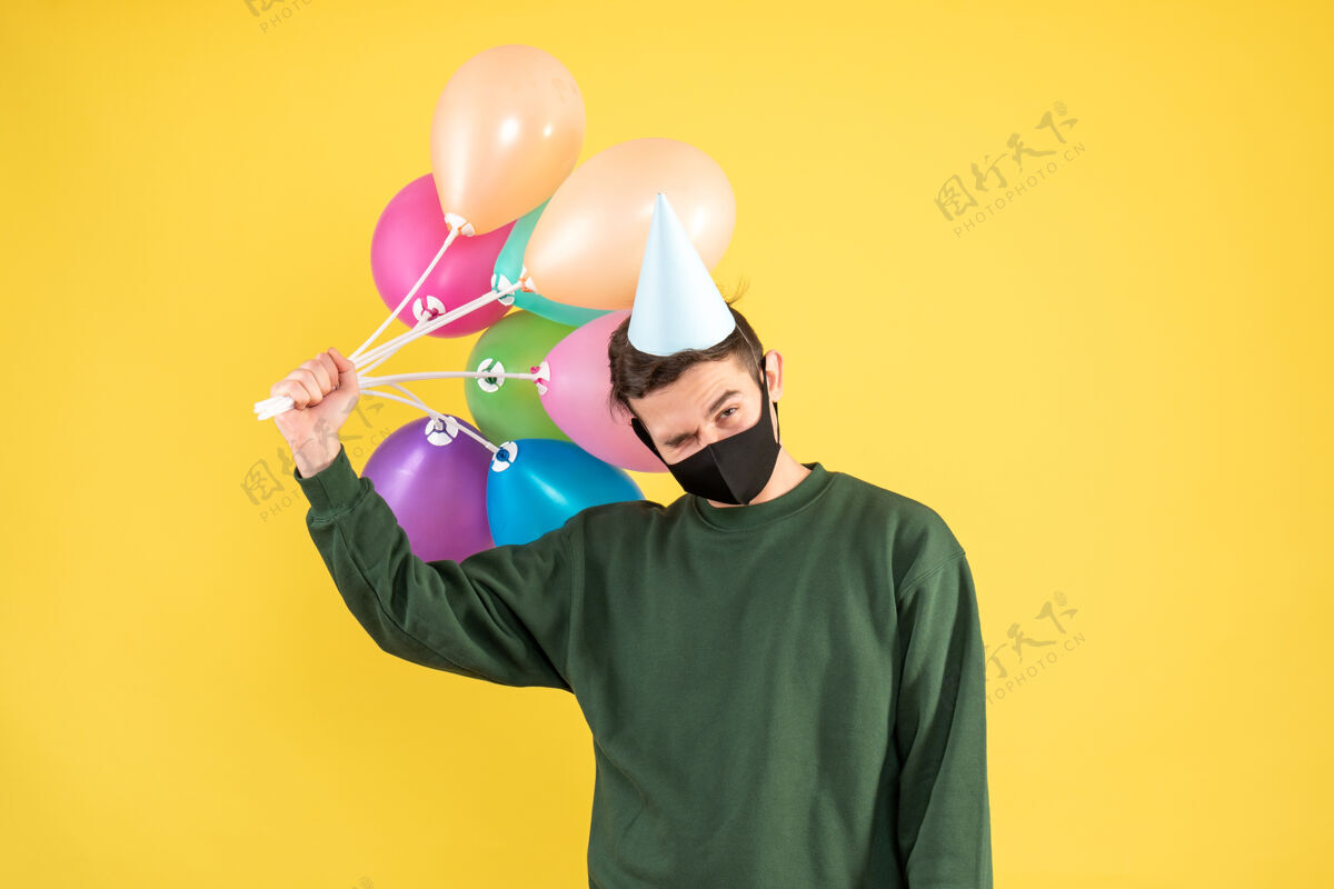 男人正面图：戴派对帽的年轻人手持五颜六色的气球站在黄色背景上卡通站立剪影