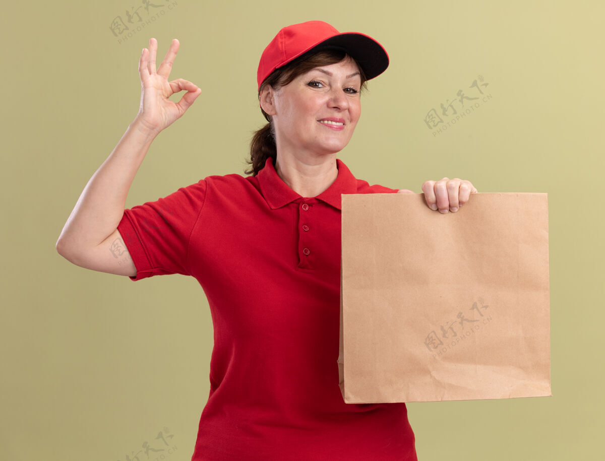 表演身穿红色制服 头戴礼帽的中年女送货员拿着纸包 面带微笑地看着前面 绿色的墙上挂着“ok”的牌子好的拿着制服