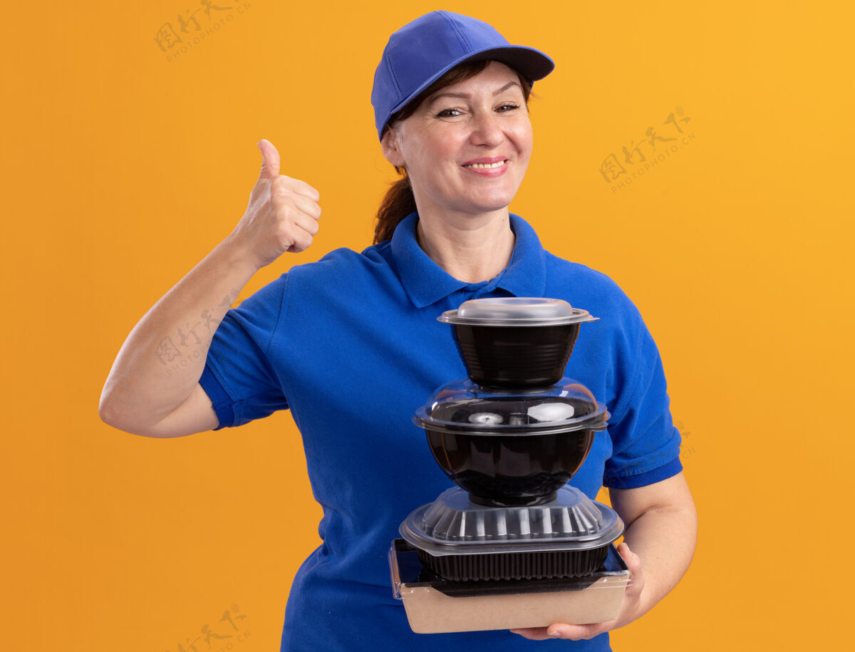 抱着快乐的中年女送货员身穿蓝色制服 戴着帽子 手里拿着一叠食品包 面带微笑 站在橙色的墙上 愉快地竖起大拇指快乐帽子食物