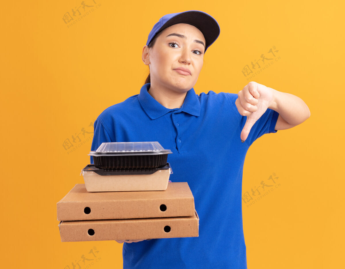 女人身穿蓝色制服 头戴鸭舌帽 拿着比萨饼盒和食品包的年轻送货员困惑地看着前面 不高兴地竖起大拇指站在橙色的墙上困惑放下站着