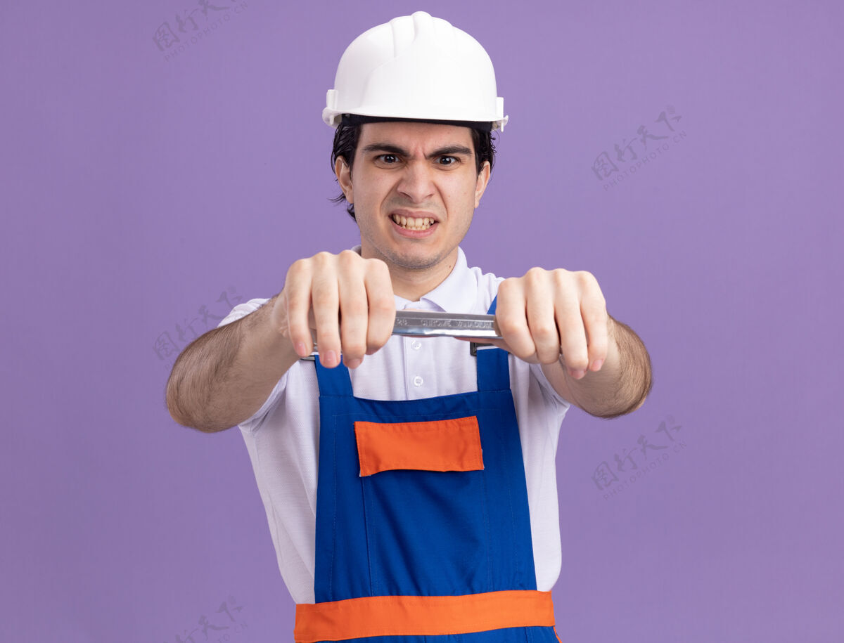 头盔身穿施工制服 头戴安全帽 手持扳手的年轻建筑工人站在紫色的墙上怒气冲冲地看着它男人站着愤怒