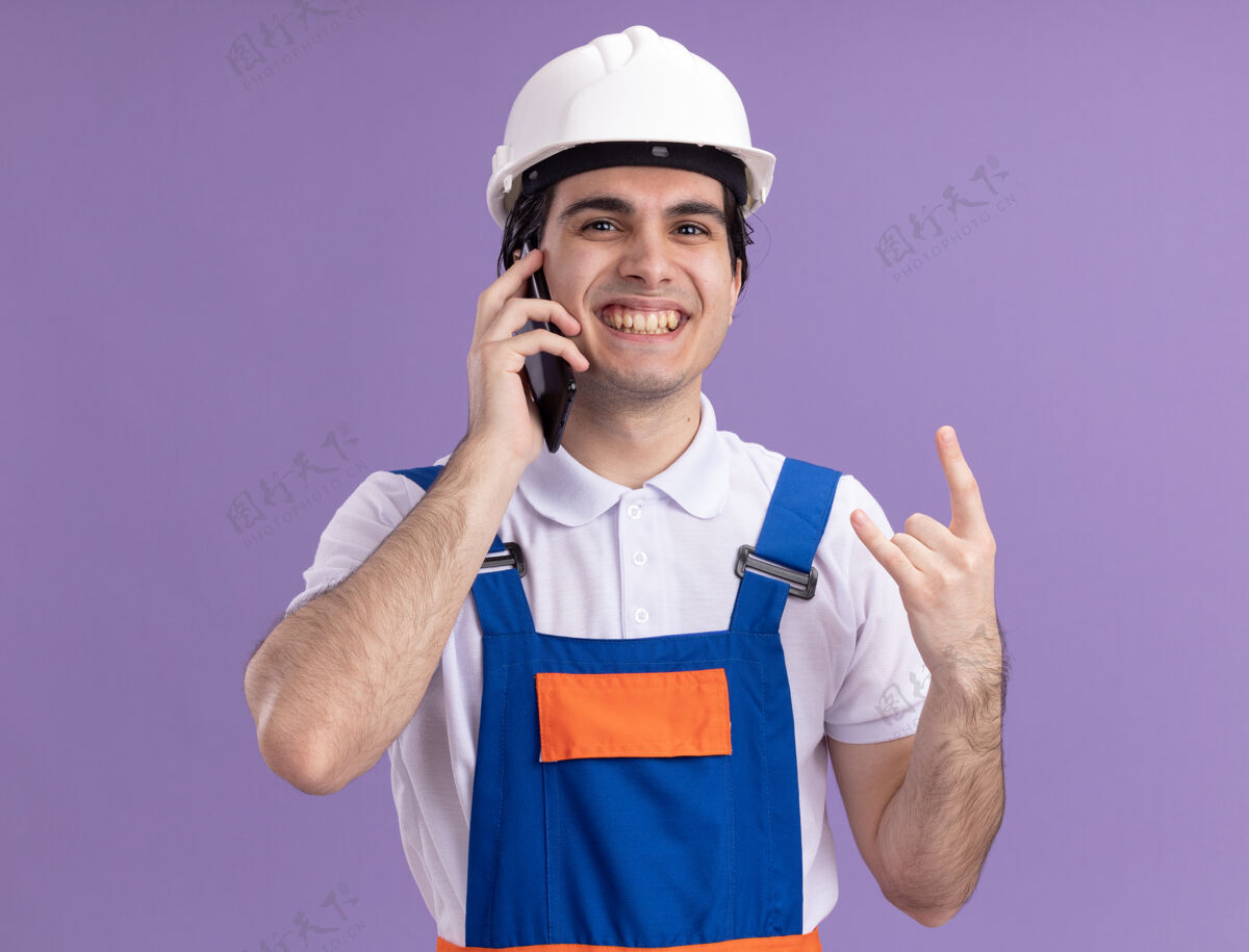 头盔身穿施工制服 头戴安全帽的年轻建筑工人站在紫色的墙上 兴高采烈地微笑着用手机制作岩石符号手机岩石站立