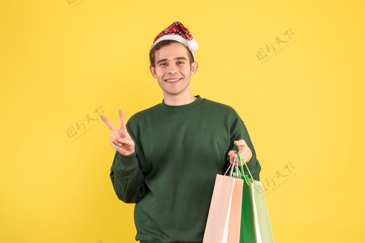 快乐正面图：戴圣诞帽的年轻人拿着购物袋在黄色背景上做胜利标志帽子男性购物