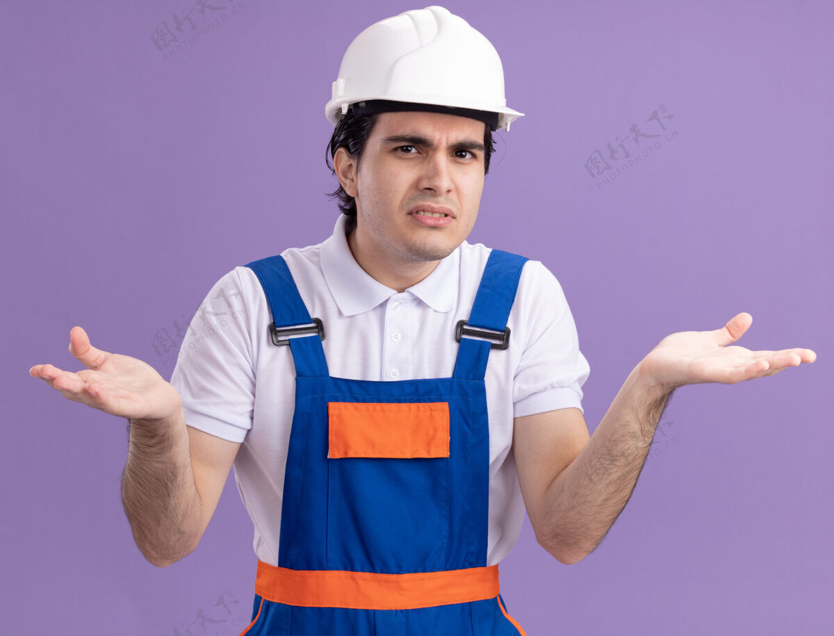 困惑身穿施工制服 头戴安全帽的年轻建筑工人站在紫色的墙上 困惑地 不悦地看着前方年轻站立制服