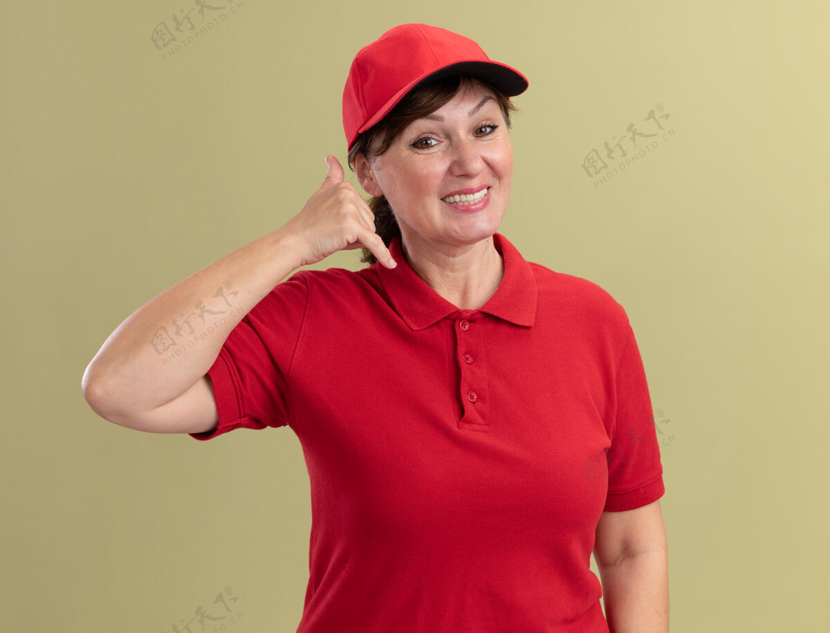 女人身穿红色制服 头戴鸭舌帽的中年女送货员站在绿色的墙边 面带微笑地打电话给我品牌中间电话