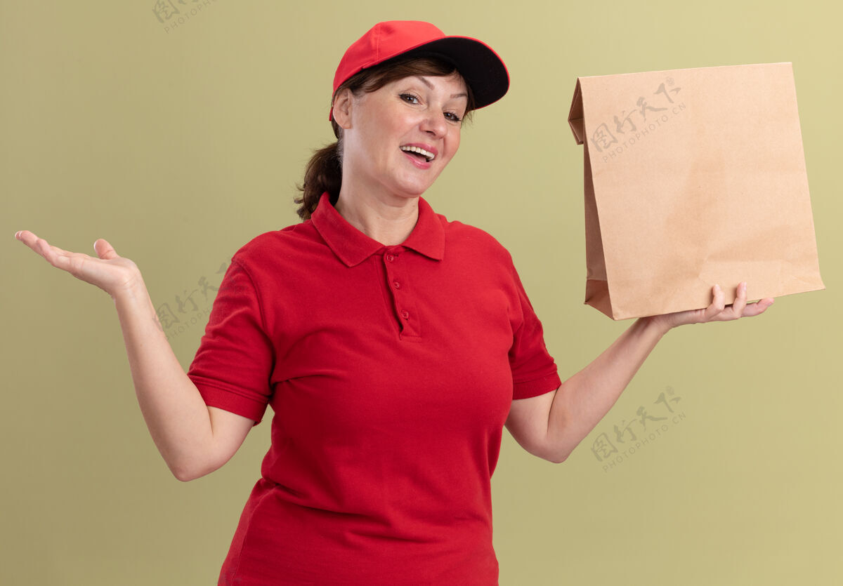 抱着快乐的中年送货员 身穿红色制服 戴着帽子 手里拿着纸包 面带微笑 站在绿色的墙上 高高兴兴地献上礼物微笑介绍手臂