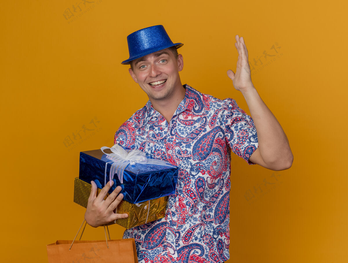 帽子快乐的年轻人戴着蓝色帽子拿着礼品盒 提着袋子 手孤立在橙色背景上礼物高兴穿