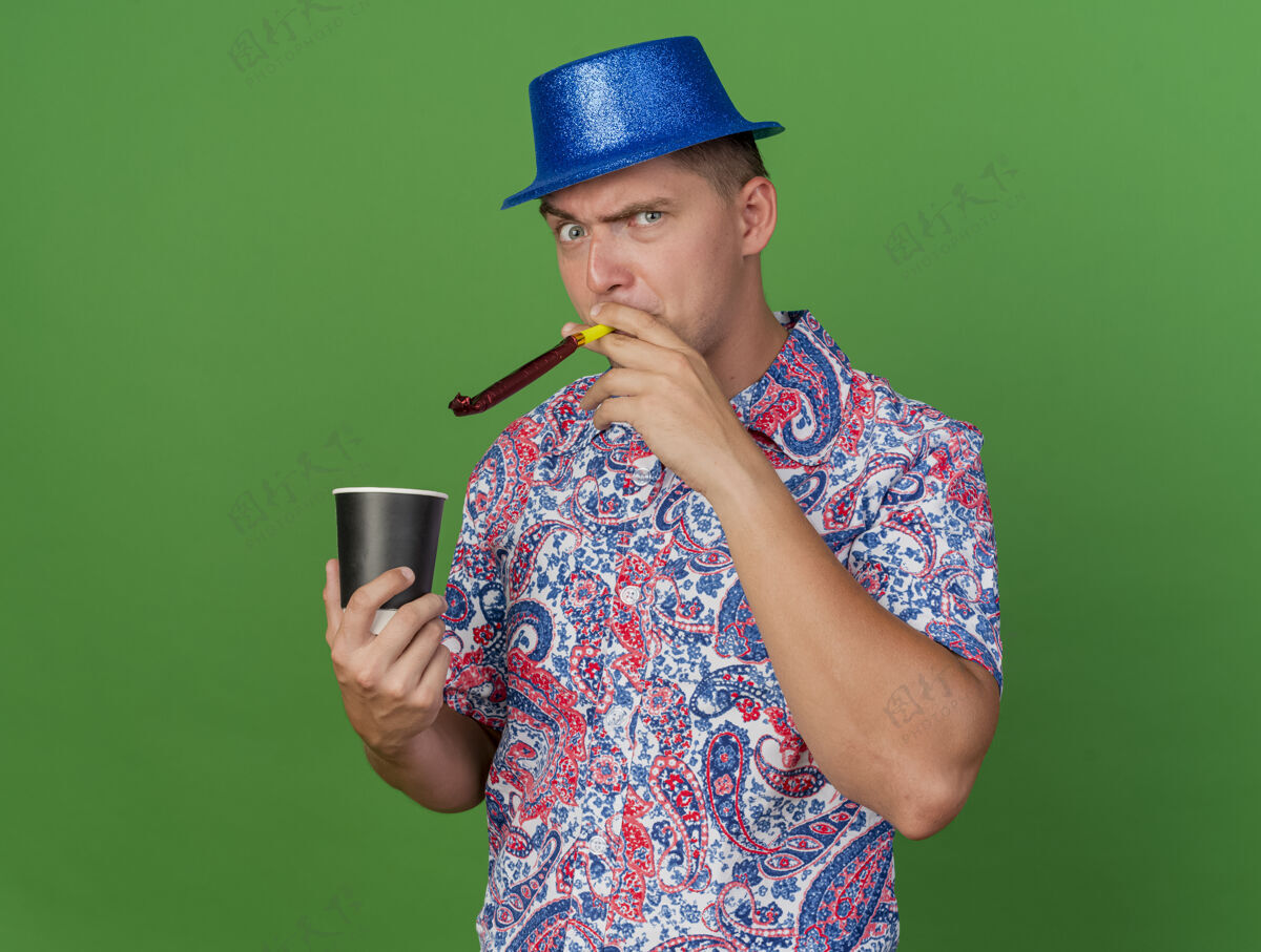 拿着愤怒的年轻人戴着蓝帽子 拿着一杯咖啡 吹着绿色背景上的派对吹风机穿杯子愤怒