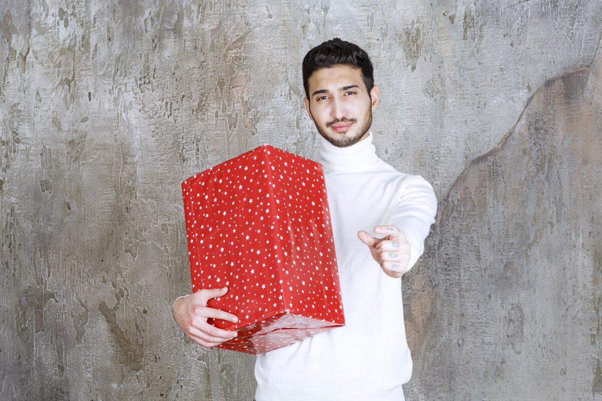 年轻一个穿白毛衣的男人手里拿着一个上面有白点的红色礼盒 并显示出积极的手势庆祝享受礼物