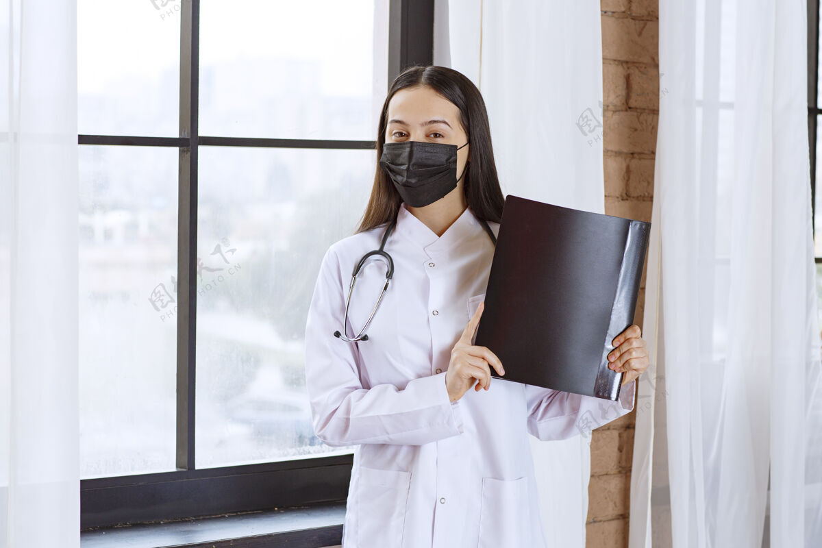 医生医生拿着听诊器和黑色口罩站在窗口旁边 手里拿着一个黑色的病人病史文件夹制服医疗诊所