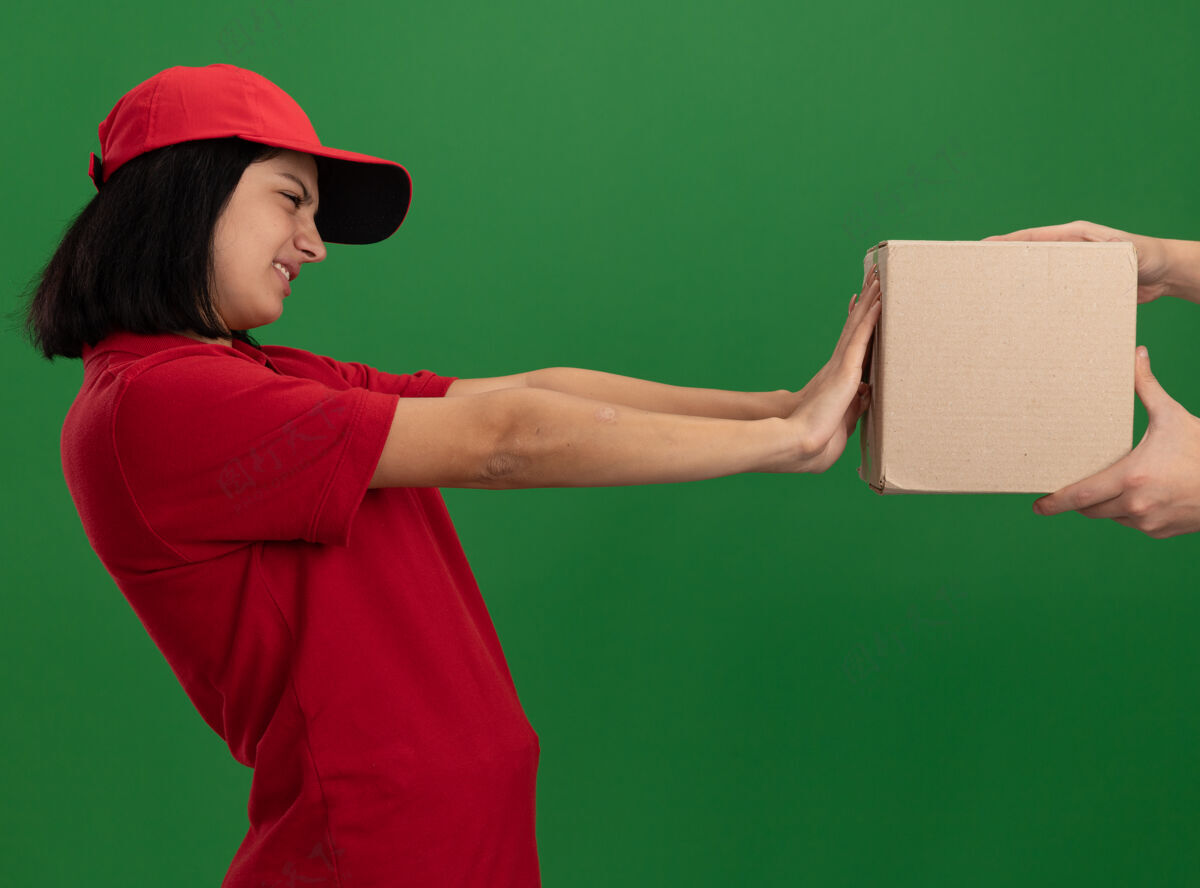 送货穿着红色制服 戴着帽子的年轻送货女孩站在绿色的墙上拒绝拿纸包裹年轻女孩帽子